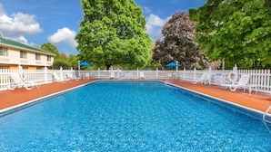 Piscina rectangular con cerramiento blanco y sillas de descanso alrededor, rodeada de árboles y un edificio con techo verde.