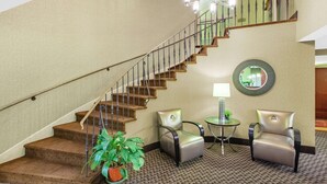 Una escalera con barandilla, una planta en maceta, dos sillones, una mesita con lámpara y un espejo en la pared.