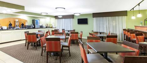 A hotel dining area with multiple tables and chairs, a kitchen area with appliances, and a large window with curtains.