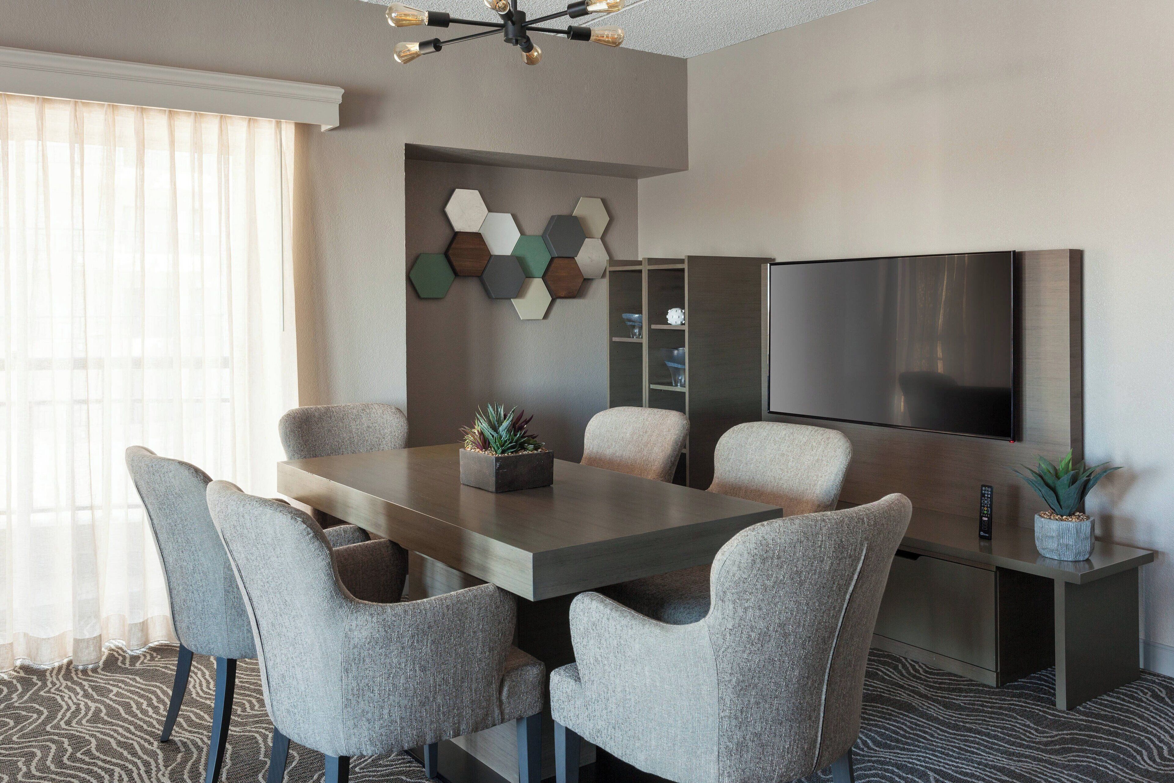 A modern dining area with a rectangular table, six grey upholstered chairs, a potted plant, and a built-in TV on the wall.