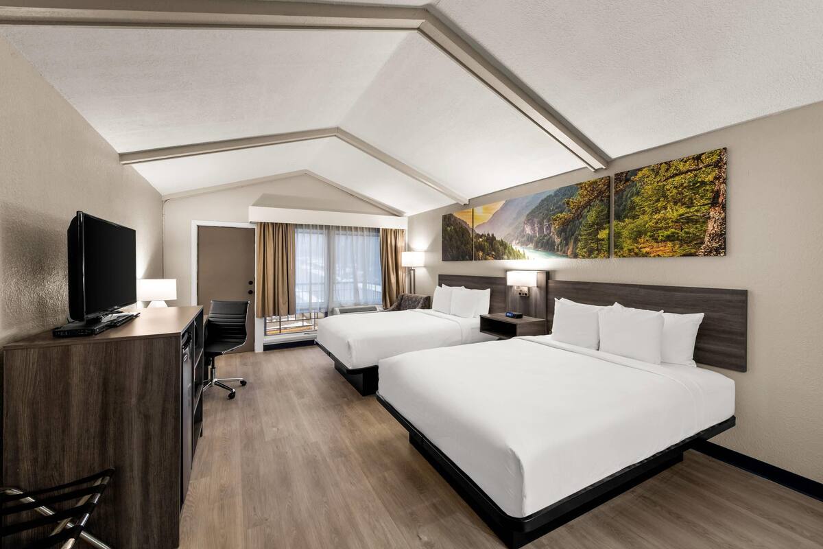 A hotel room with two beds, a desk with a TV, a chair, a desk lamp, and a window with curtains.