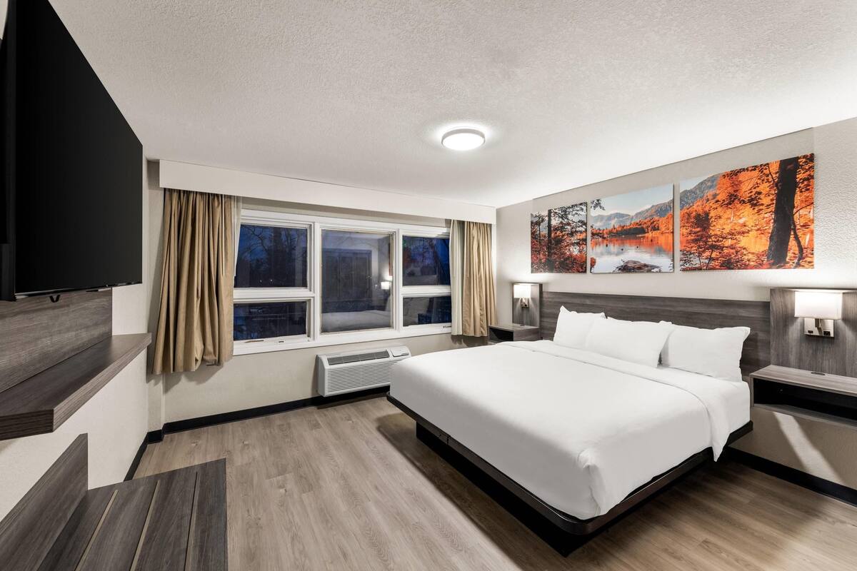 A modern hotel room with a large bed, a flat-screen TV, a window with curtains, and a wall-mounted artwork.