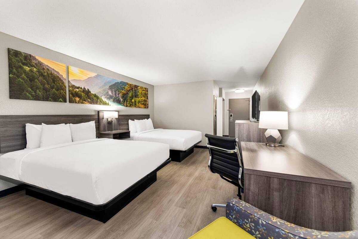 A hotel room with two beds, a desk with a chair, a mirror, and a painting of a mountain landscape.