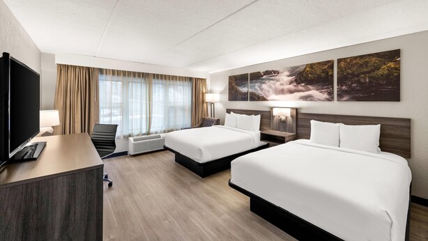 A hotel room with two beds, a desk, a TV, and a large window with curtains.