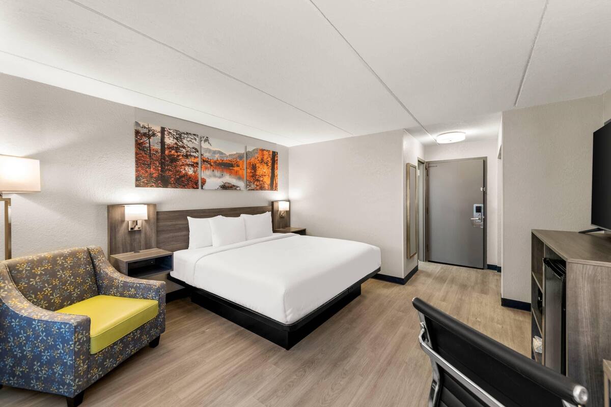 A hotel room with a bed, a sofa, a desk, a TV, and artwork on the wall.