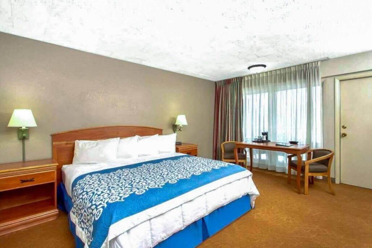 A hotel room with a bed, a desk, a chair, a lamp, and a door.