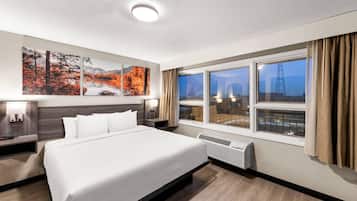 A hotel room with a large bed, a nightstand, a wall-mounted lamp, a window with curtains, and a view of a cityscape.