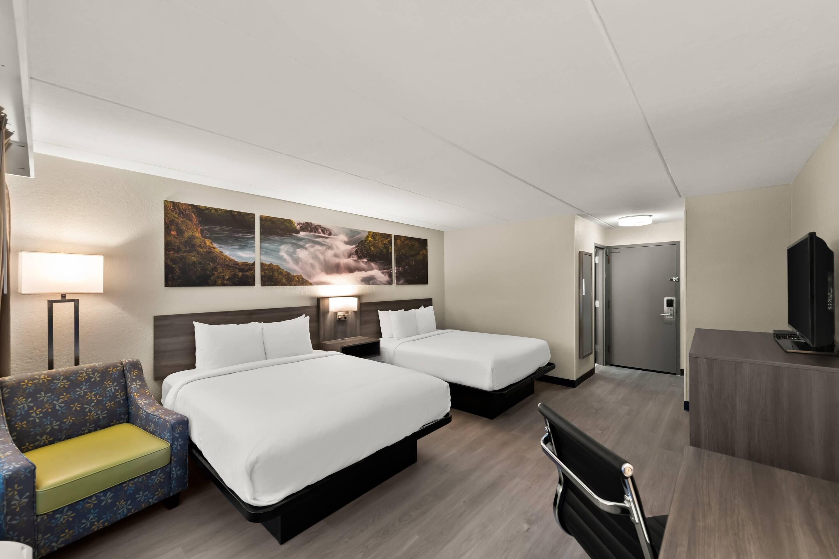 A hotel room with two beds, a sofa, a desk, a TV, and artwork on the wall.