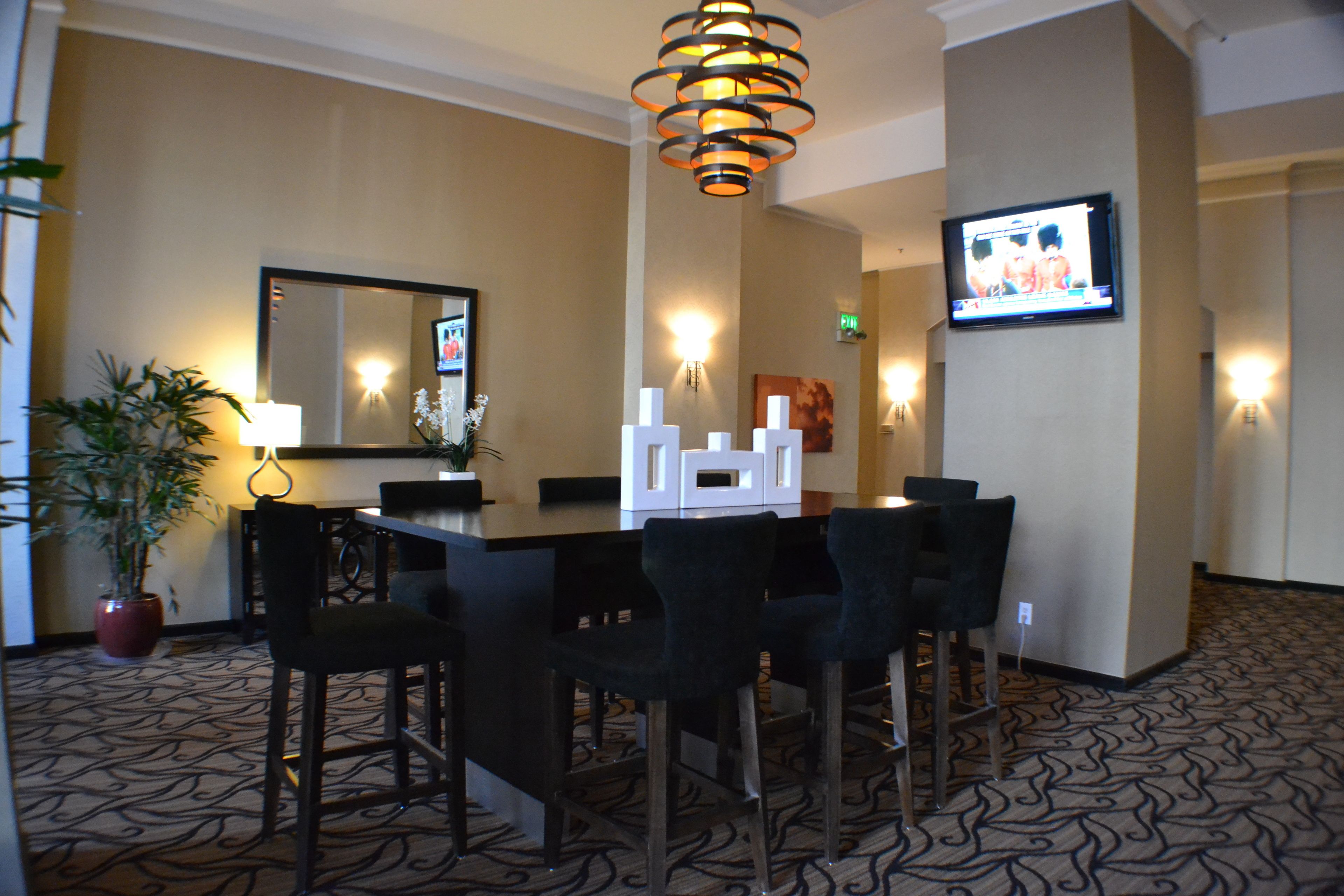 A hotel lobby with a reception desk, chairs, a potted plant, a large mirror, wall sconces, and a television mounted on the wall.