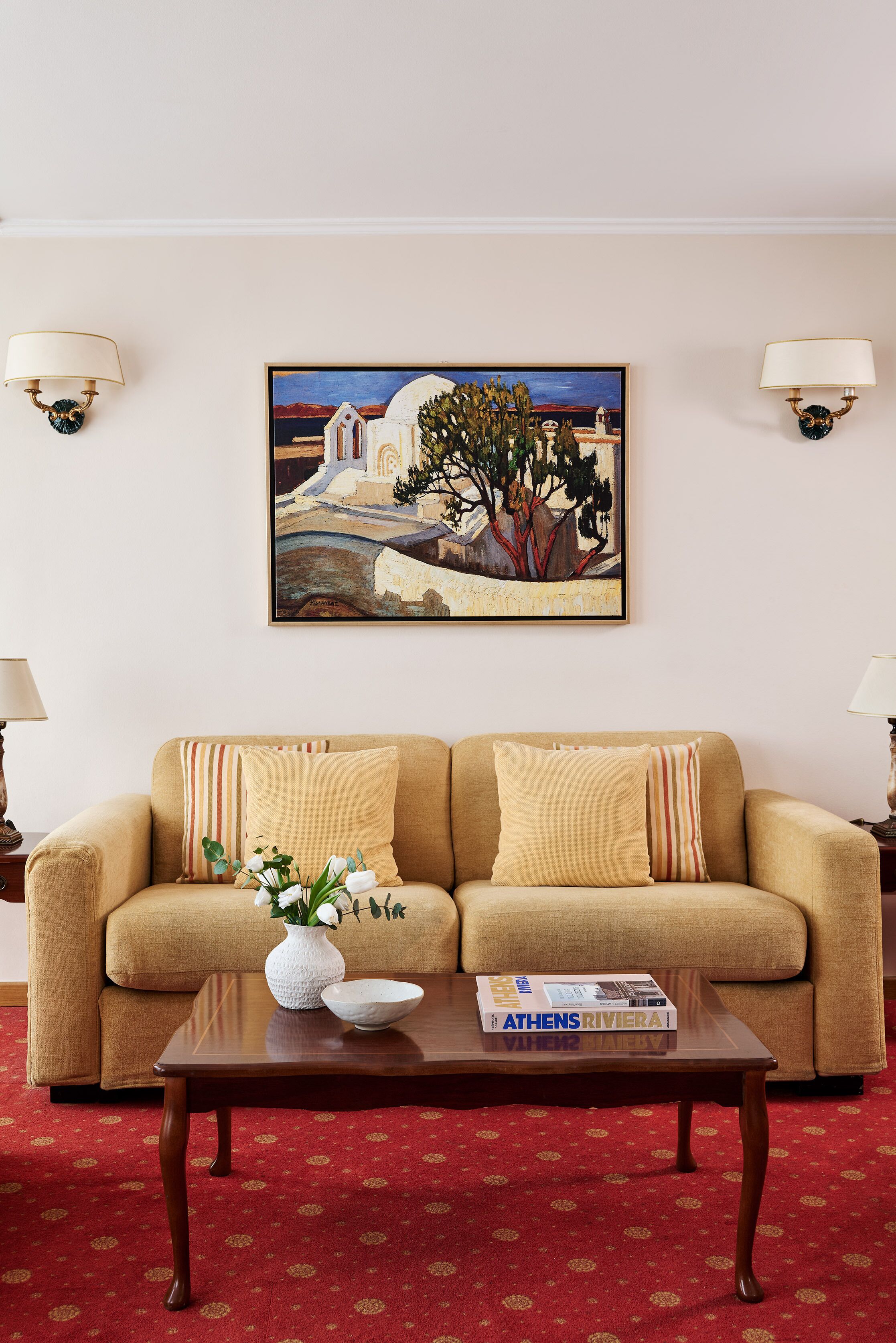 Una sala de estar con un sofá beige, una mesa de centro con un jarrón y un libro, y un cuadro de un paisaje en la pared.