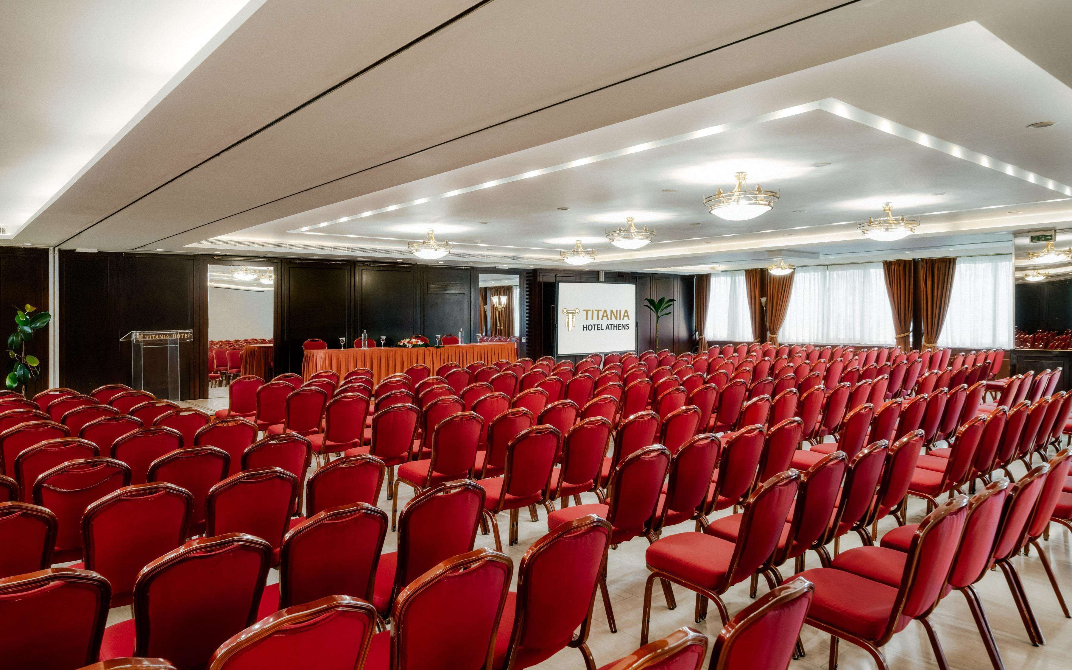 A conference room with rows of red chairs, a podium, and a screen displaying 'TITANIA HOTEL ATHENS'.