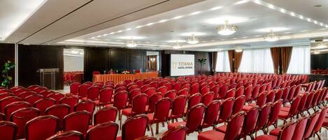 A conference room with rows of red chairs, a podium, and a screen displaying 'TITANIA HOTEL ATHENS'.