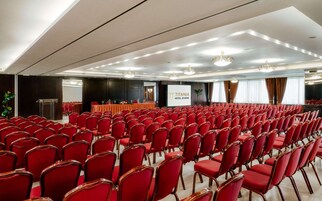 Μια αίθουσα συνεδριάσεων με σειρές από κόκκινες καρέκλες, ένα βήμα και μια οθόνη που προβάλλει την ένδειξη «ΞΕΝΟΔΟΧΕΙΟ ΤΙΤΑΝΙΑ, ΑΘΗΝΑ».