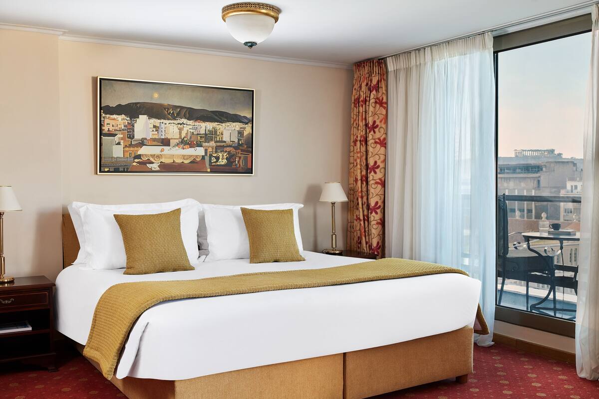 Habitación de hotel con una cama grande, dos mesitas de noche, una mesita auxiliar, un cuadro en la pared y vista a la ciudad a través de un ventanal amplio.