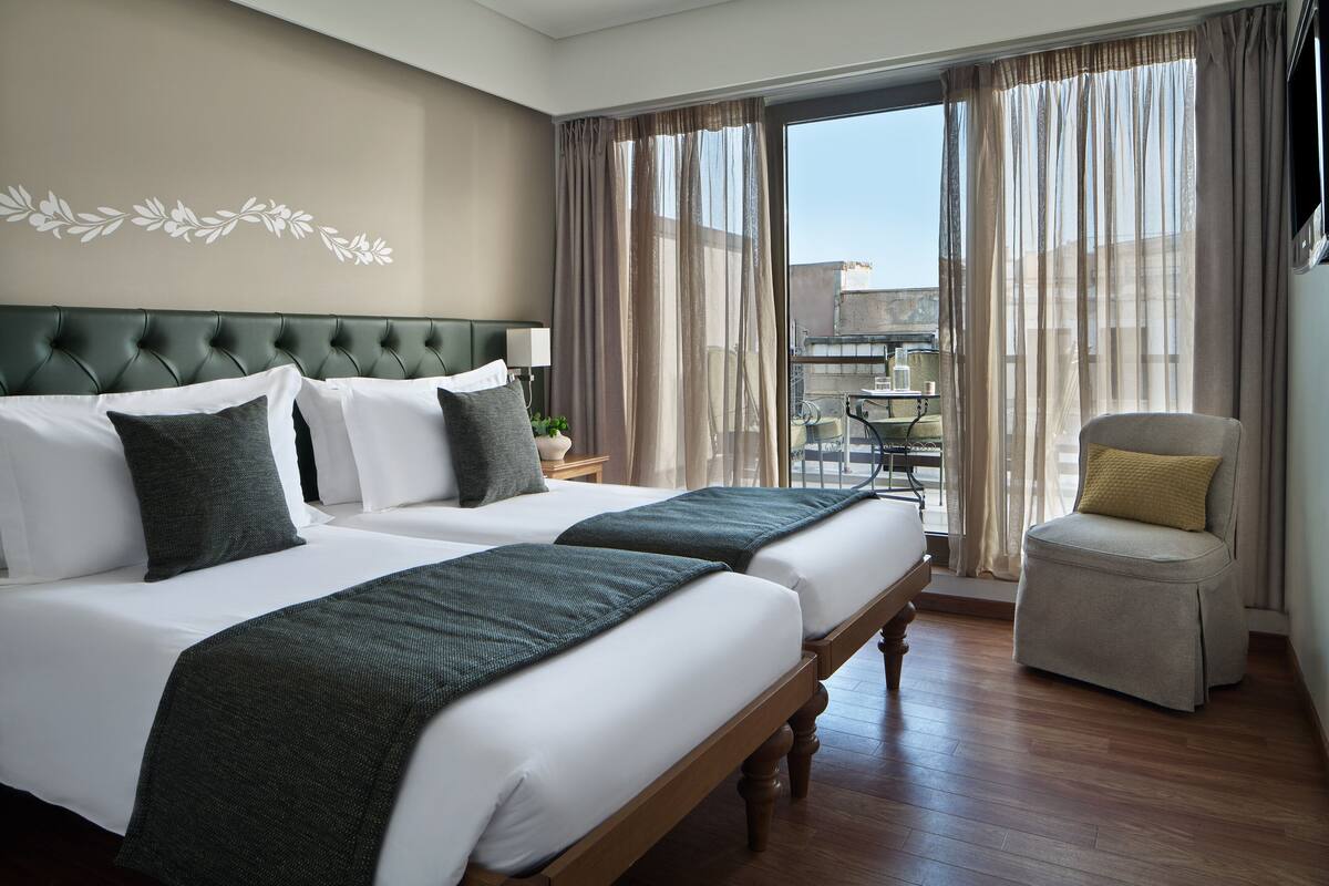 Habitación de hotel con dos camas, un cabecero tapizado en verde, una mesita de noche con lámpara y vistas al exterior a través de cortinas traslúcidas.