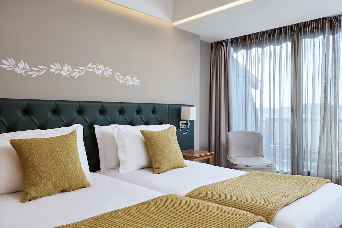 Habitación de hotel con dos camas, cabecera tapizada de color verde, ropa de cama blanca y vistas de la ciudad a través de cortinas traslúcidas.