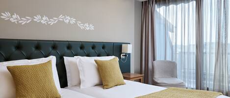 A hotel room with two beds, a green upholstered headboard, white bedding, and a view of the city through sheer curtains.