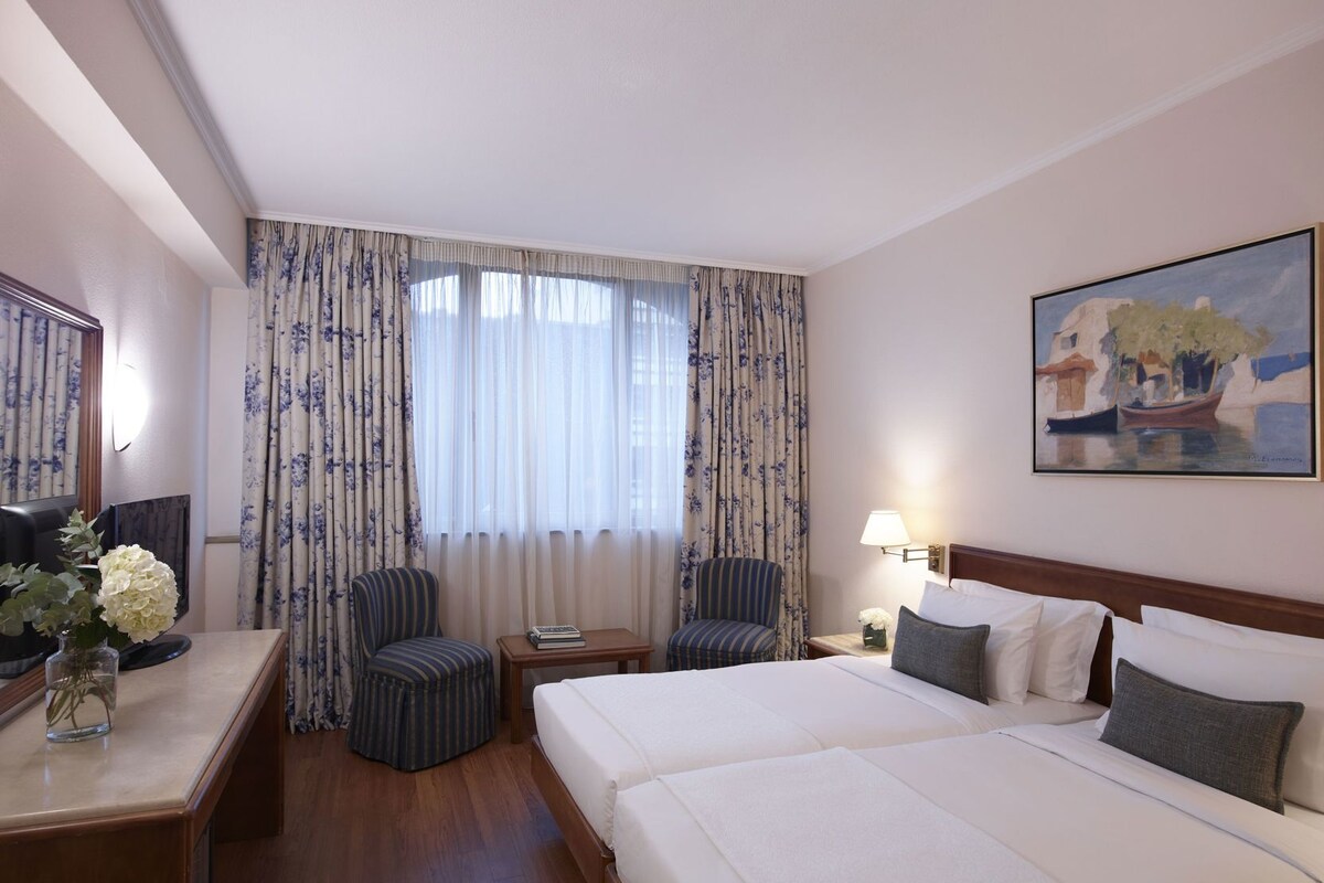 Quarto de hotel com cama, duas cadeiras, escrivaninha, abajur, pintura e janela com cortinas.