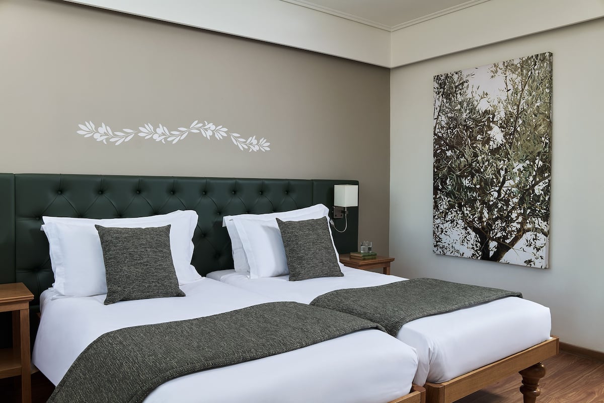 Un dormitorio con cabecera capitonada, dos camas con sábanas blancas y mantas de color verde oscuro, una mesita de noche con lámpara y un cuadro colgado en la pared con un árbol.
