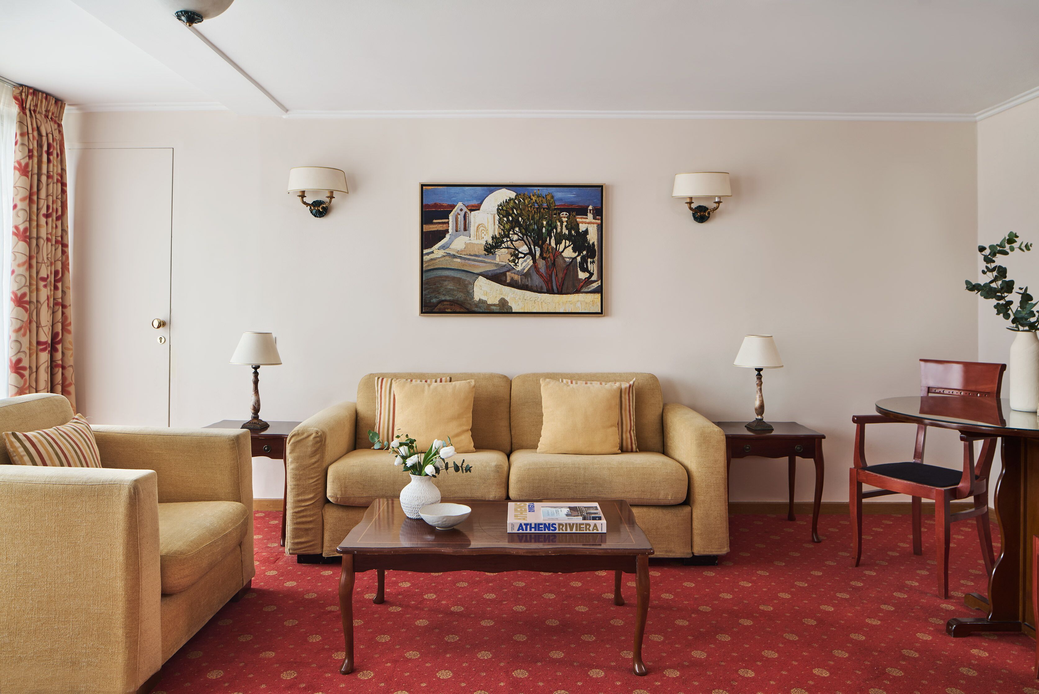 Una sala de estar con un sofá color beige, una mesa de centro de madera, una mesita auxiliar, una silla y un cuadro en la pared.
