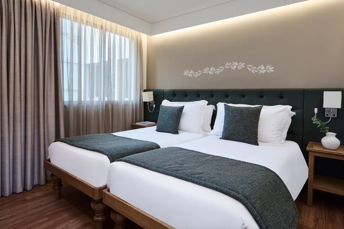 Habitación de hotel con dos camas, un ventanal con cortinas, una lámpara de pared y una mesita de noche con un florero.
