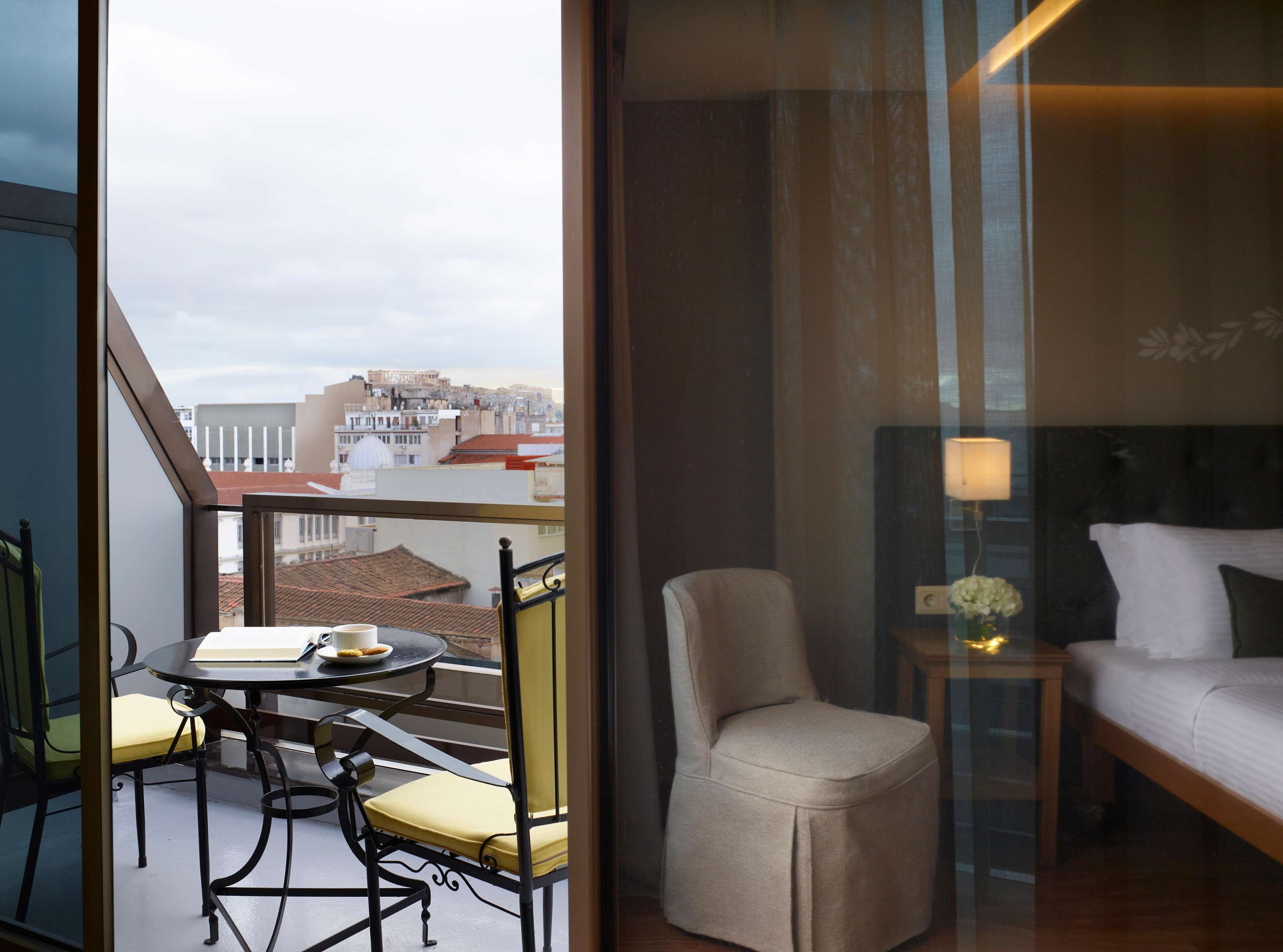 A hotel room with a balcony, a bed, a chair, a table with a book and a cup, and a view of the cityscape.