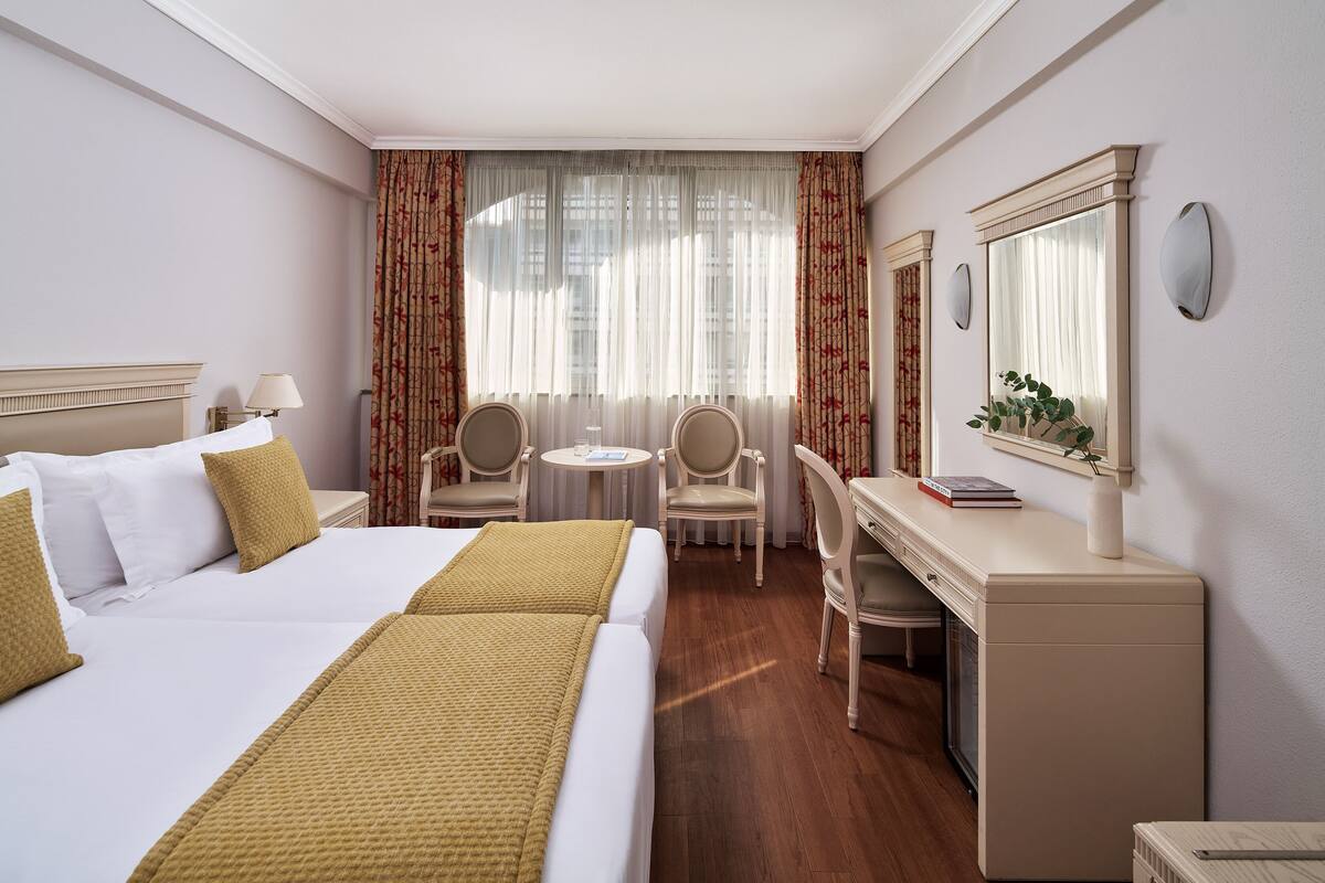 Habitación de hotel con cama, escritorio, dos sillas, una mesita, un espejo y ventana con cortinas.