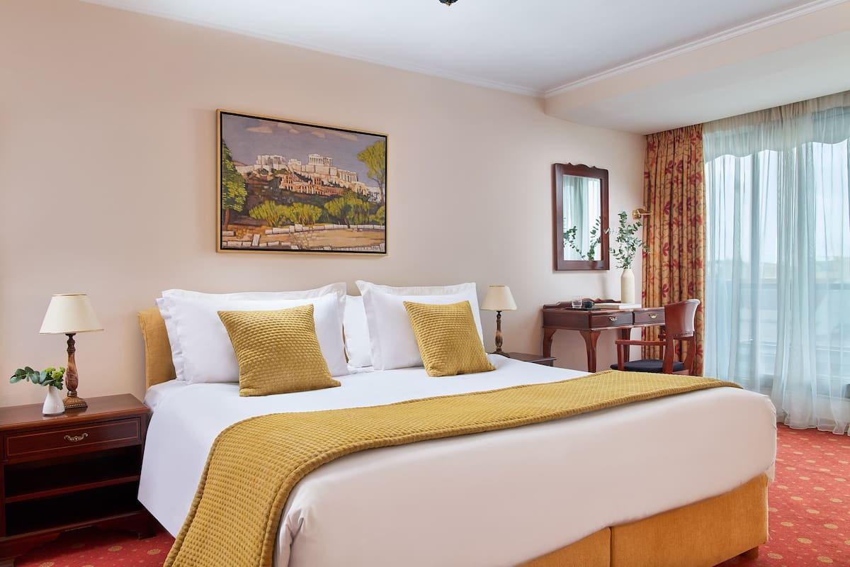 Habitación de hotel con una cama grande, un escritorio con silla, una mesita de noche, una lámpara, un espejo y un cuadro de un paisaje urbano.