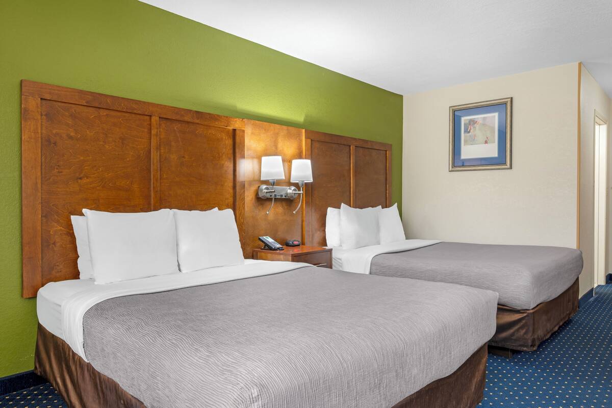 A hotel room with two beds, a wooden headboard, a nightstand with a lamp, a framed picture on the wall, and a blue carpet.