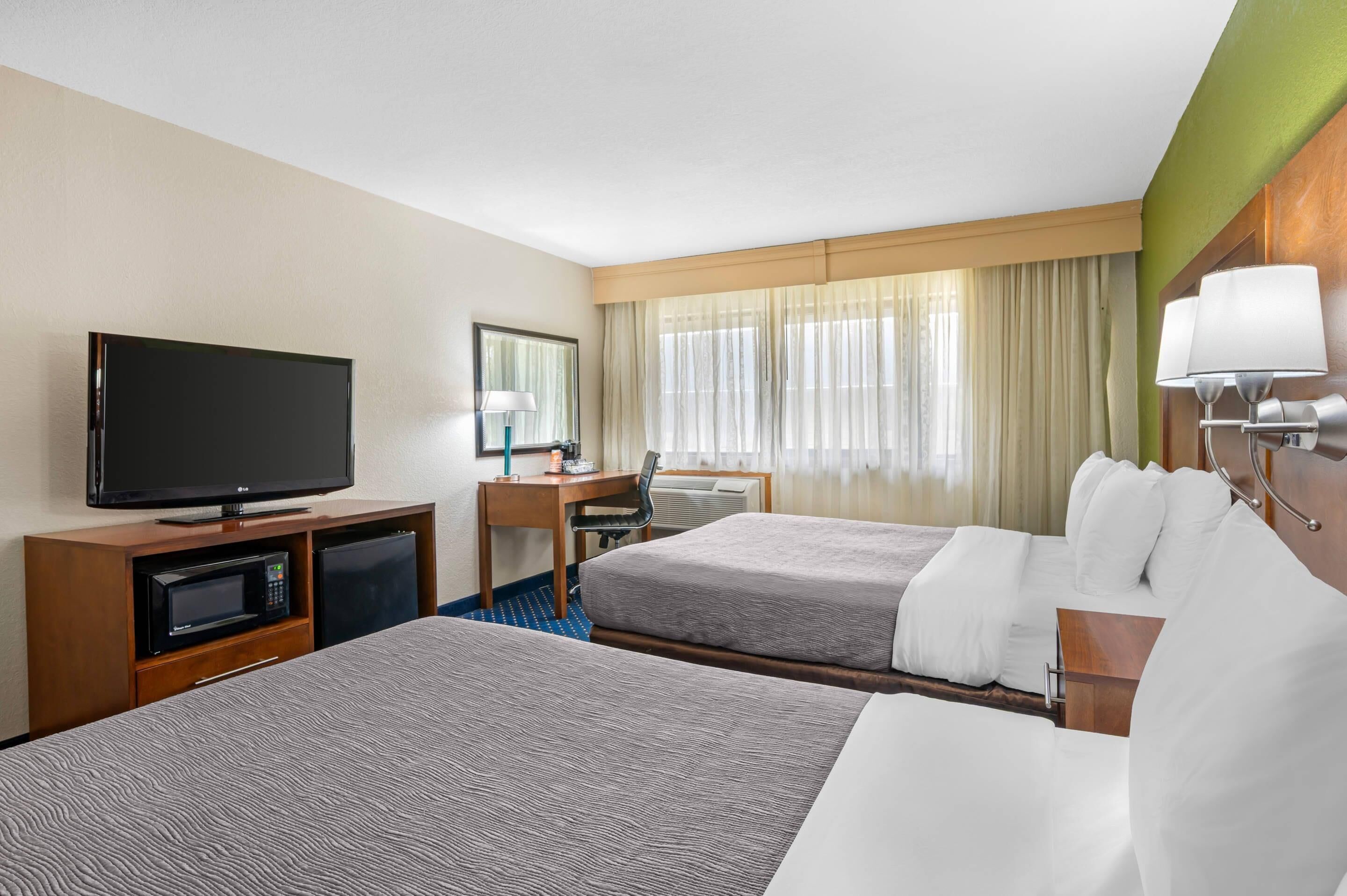 A hotel room with two beds, a TV, a desk with a chair, a microwave, and a window with curtains.