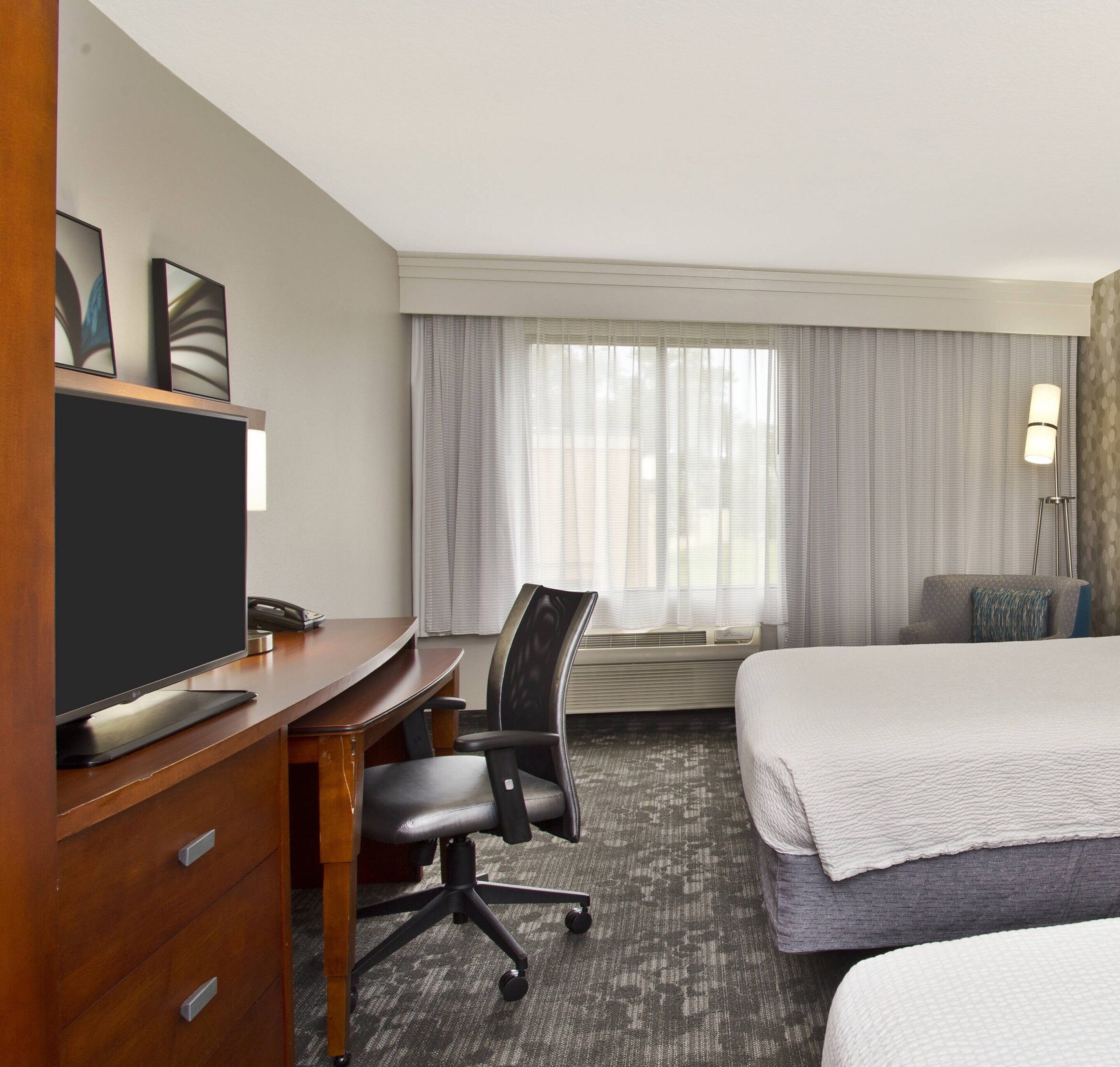 Une chambre d’hôtel comprenant un lit, un bureau avec un ordinateur, une chaise, une télévision, une fenêtre avec des rideaux et une lampe.