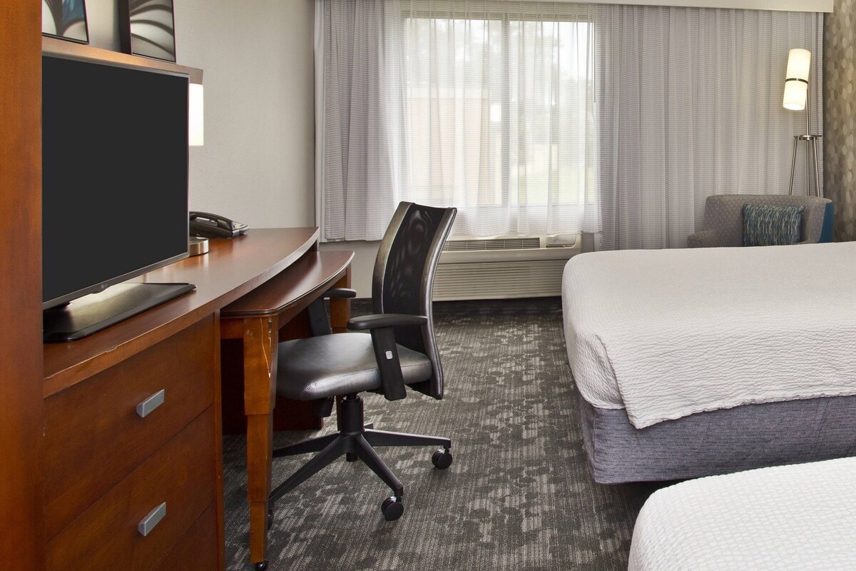 A hotel room with a bed, a desk with a computer, a chair, a TV, a window with curtains, and a lamp.