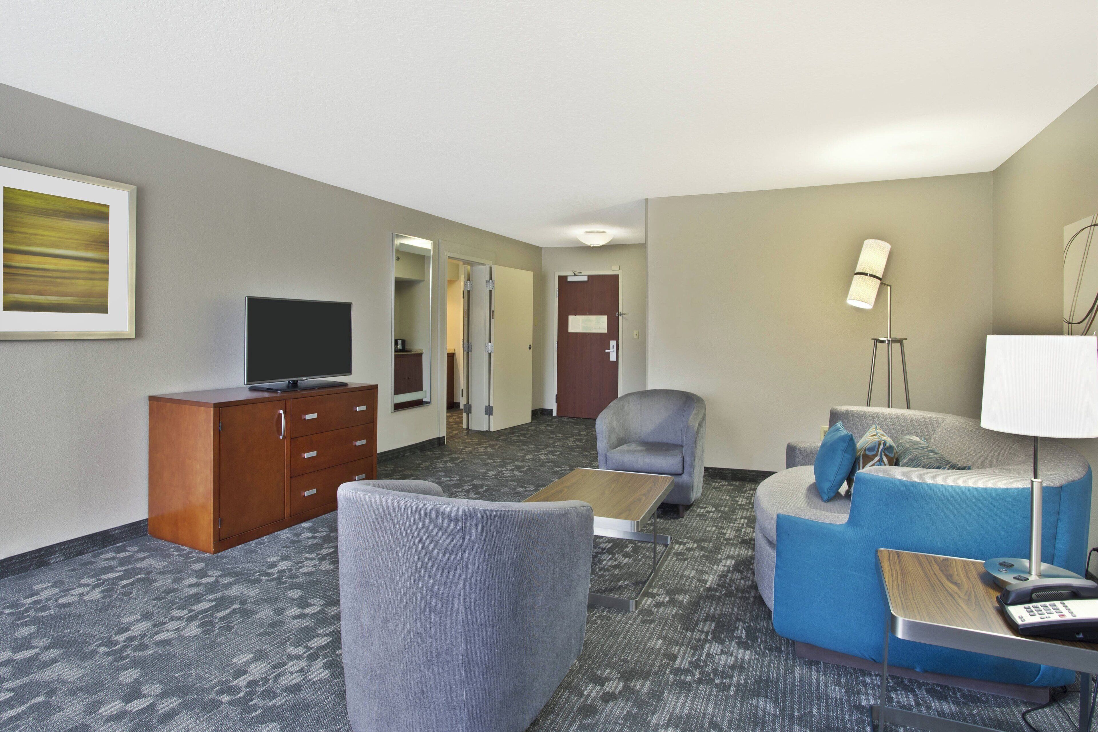 A hotel room with a flat-screen TV, a wooden cabinet, a grey armchair, a blue sofa, a small table, and a lamp.