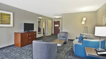 A hotel room with a flat-screen TV, a wooden cabinet, a grey armchair, a blue sofa, a small table, and a lamp.