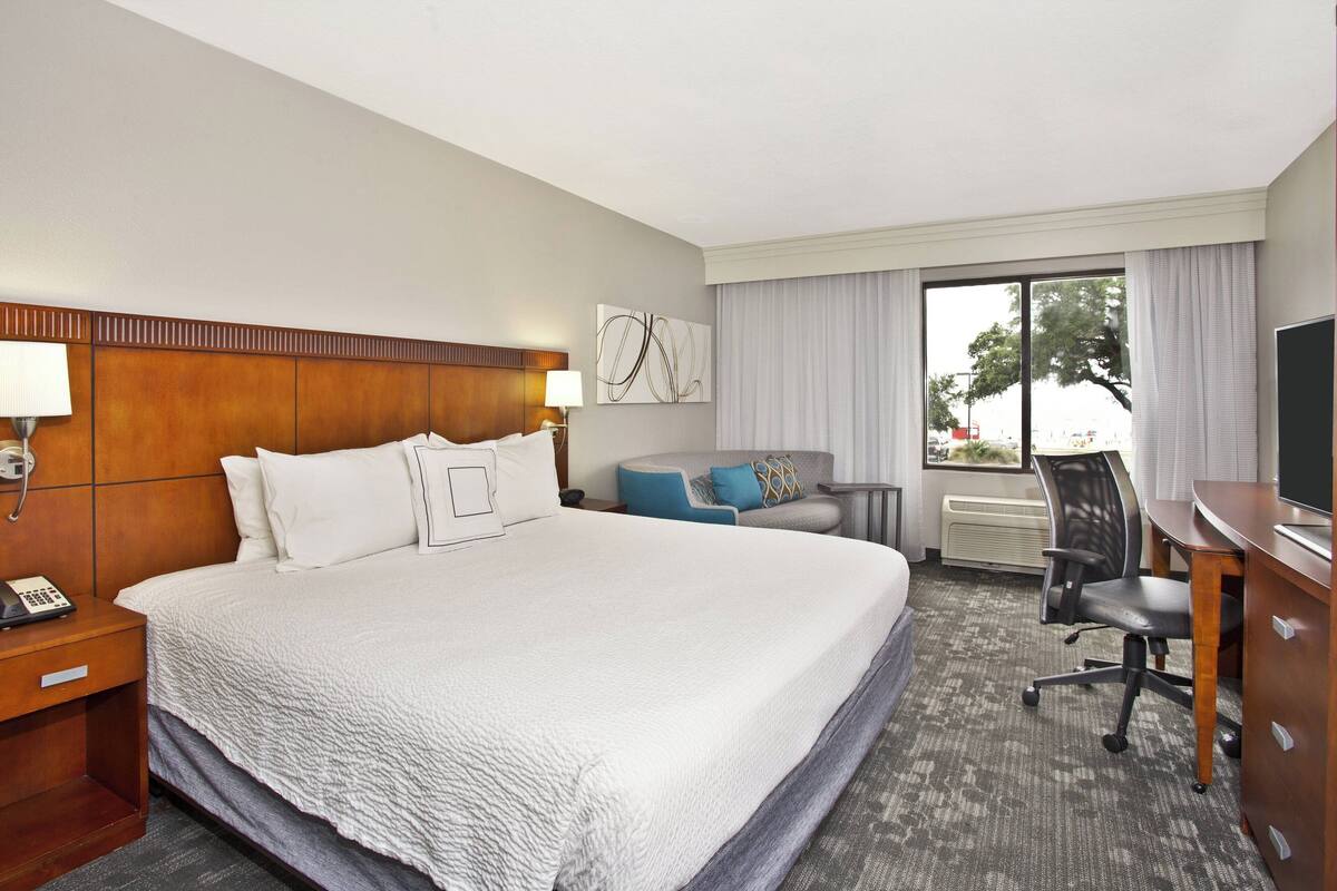 A hotel room with a large bed, a desk, a chair, a lamp, and a window with curtains.