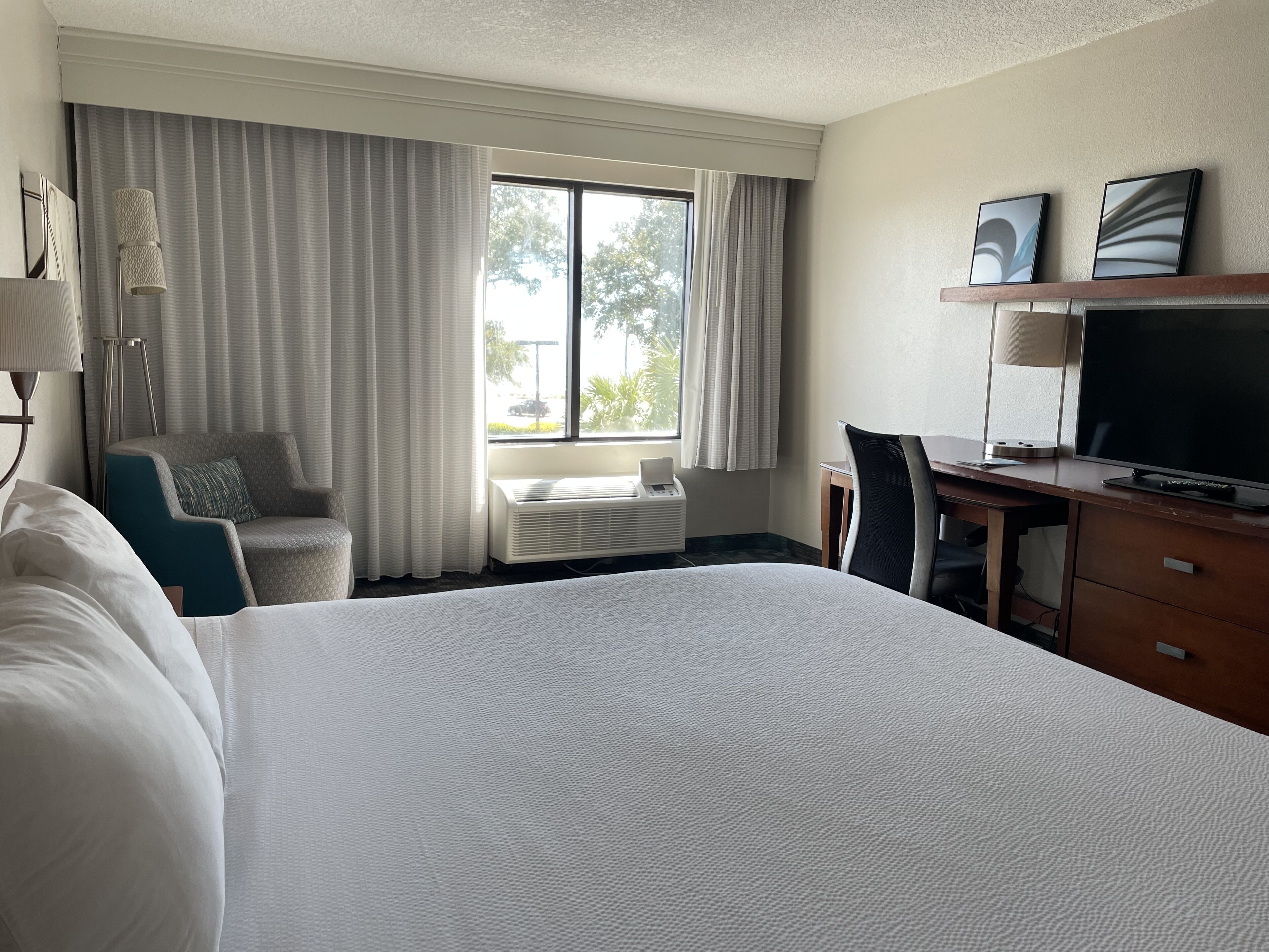 A hotel room with a large bed, a desk with a TV, a chair, a desk lamp, and a window with curtains.