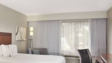 A hotel room with a large bed, a desk, a chair, a lamp, and a window with sheer curtains.