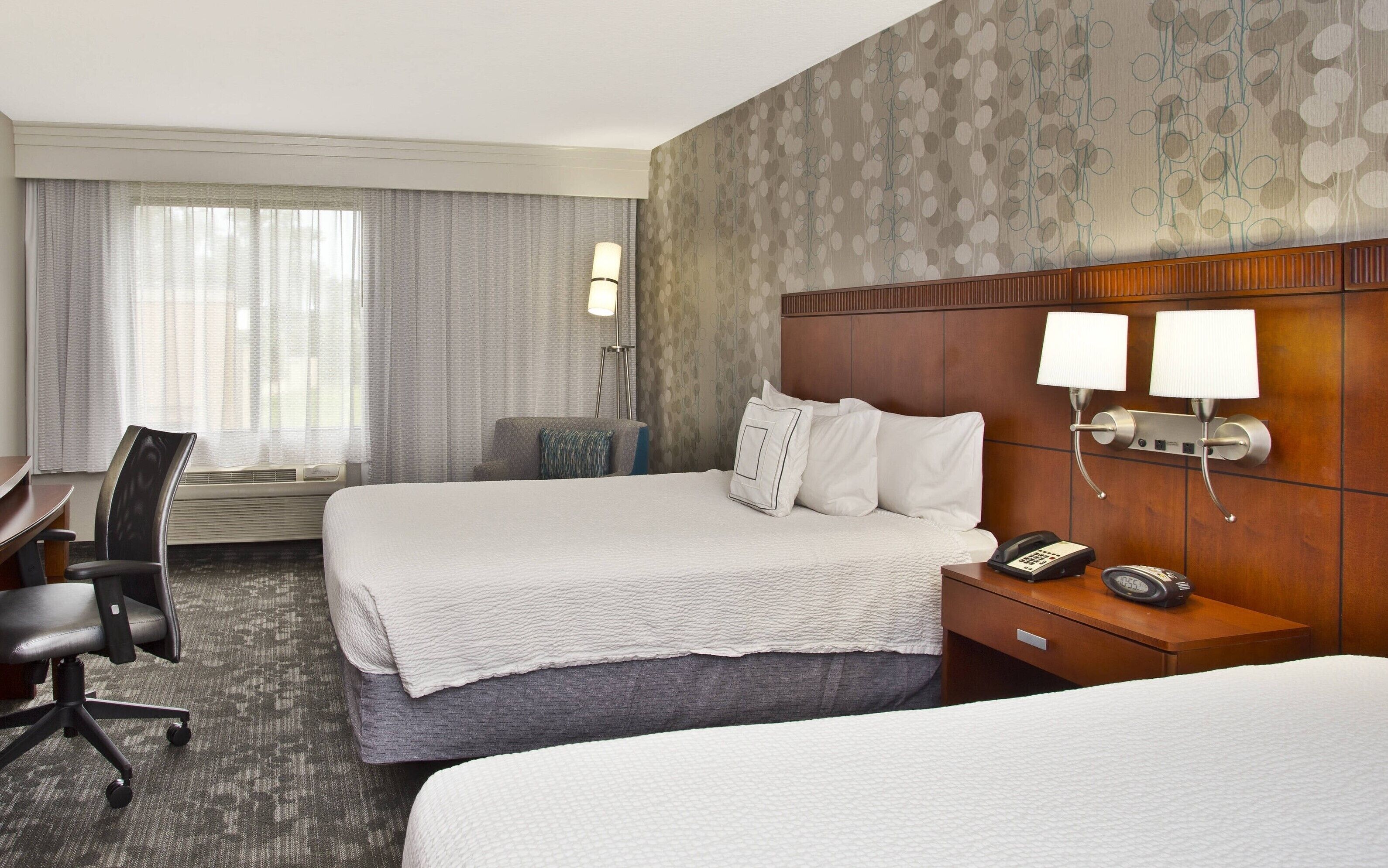 A hotel room with two beds, a desk, and a chair. There are two bedside lamps, a phone, and a clock on the nightstand.