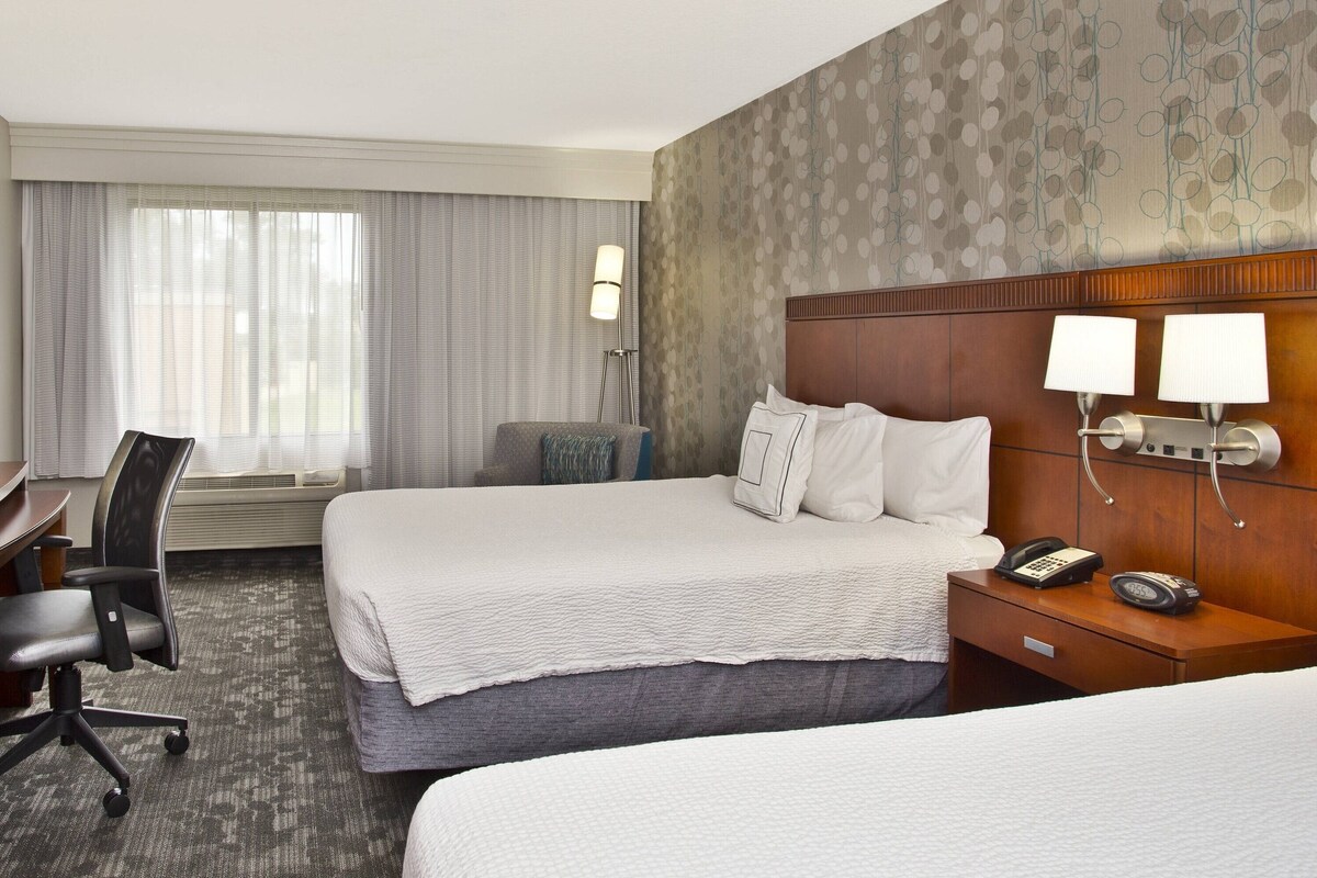 A hotel room with two beds, a desk, and a chair. There are two bedside lamps, a phone, and a clock on the nightstand.