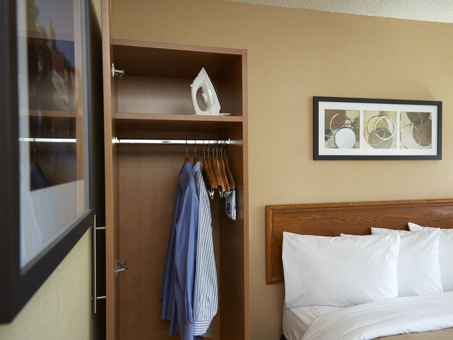 Habitación de hotel con cabecera de madera, cama con ropa blanca, armario con prendas colgadas y un cuadro abstracto enmarcado en la pared.