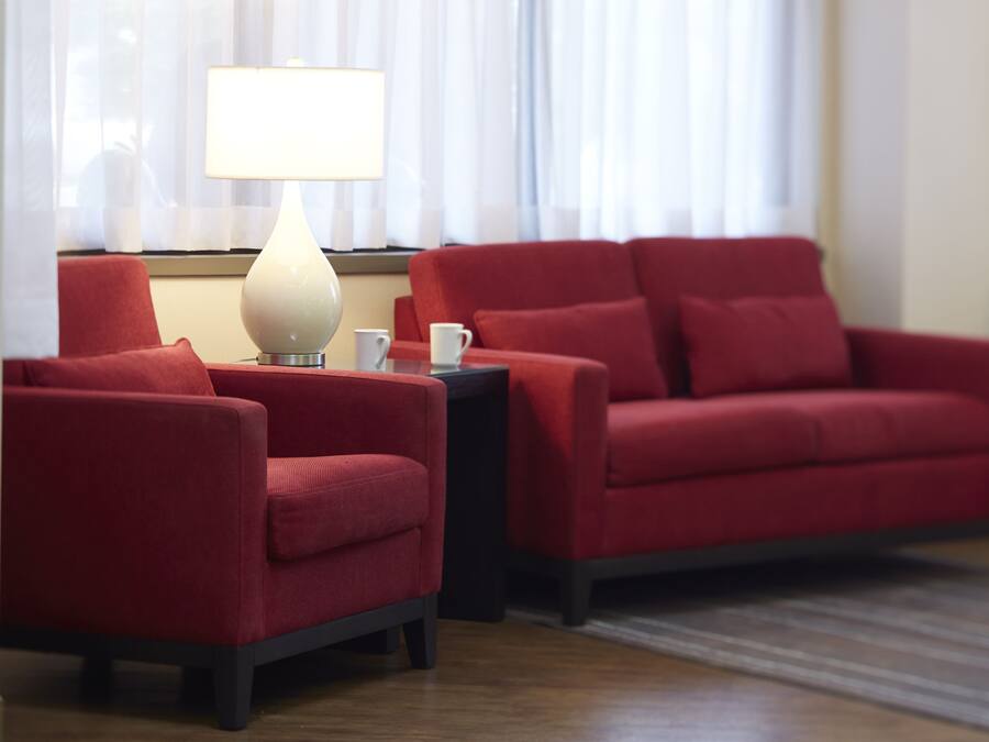 Een kamer met een rode bank, een kleinere fauteuil, een bijzettafeltje met een lamp en een raam met dunne gordijnen.
