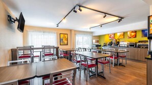 Un comedor moderno con varias mesas y sillas, una zona de cocina con equipo de cocina, y un televisor montado en la pared.
