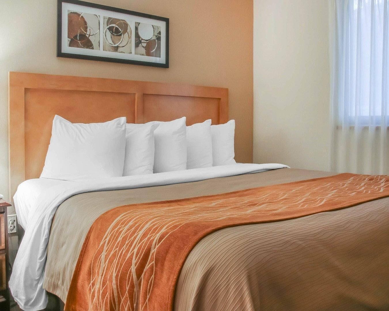 Une chambre d’hôtel avec un lit, une tête de lit, un tableau encadré au mur et une fenêtre avec des rideaux.