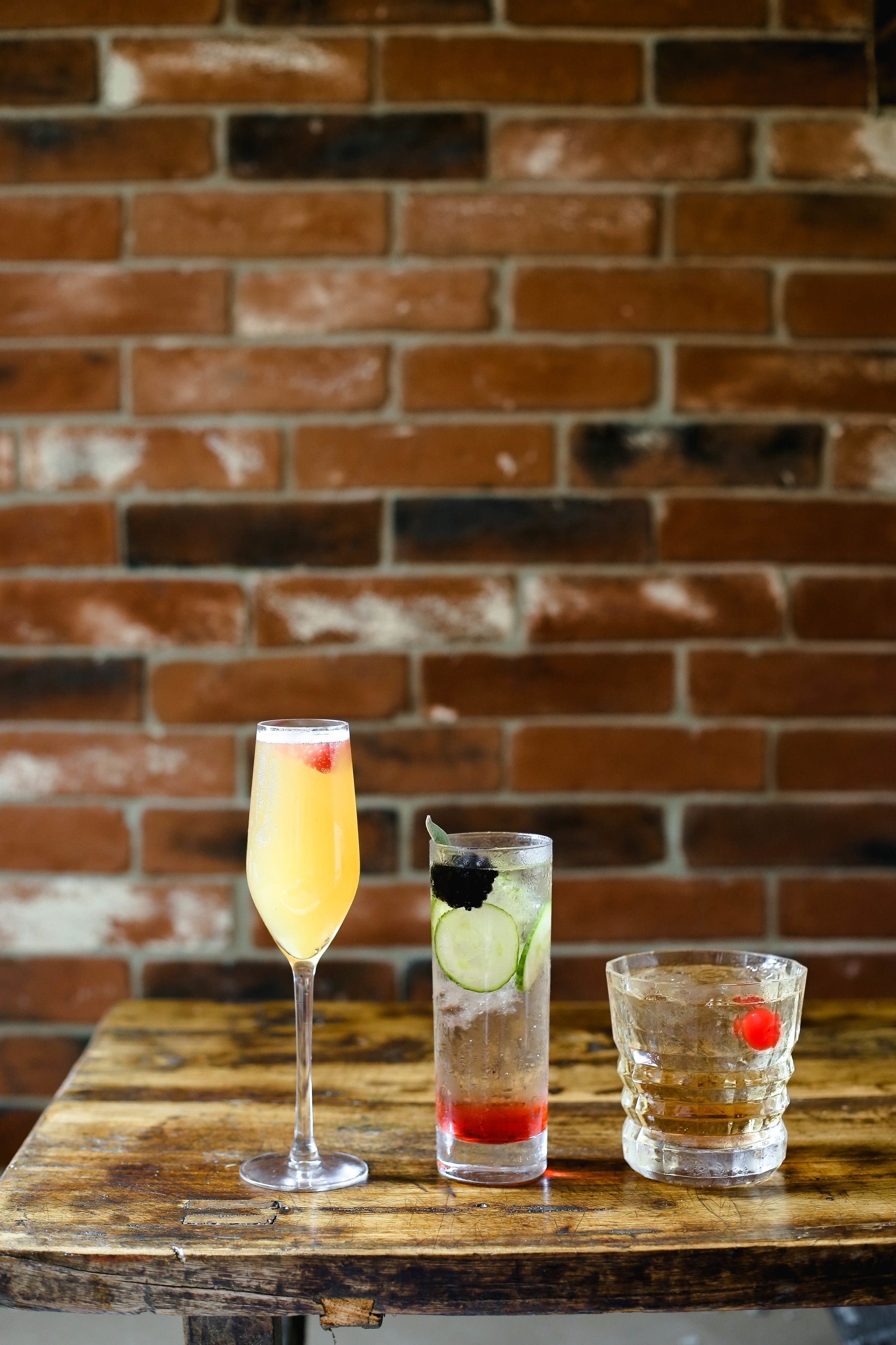 Three glasses of beverages on a wooden table against a brick wall.