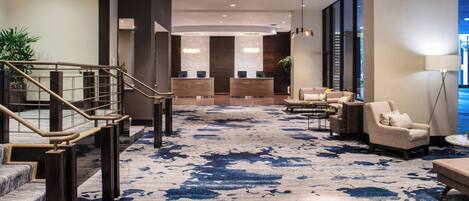 Un vestíbulo de hotel con alfombra de diseño azul, un mostrador de recepción, una zona de estar y una escalera.