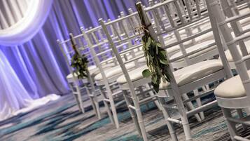 Rows of white chairs with greenery on the backrests, arranged in a formal setting with a blue carpet and draped white fabric in the background.