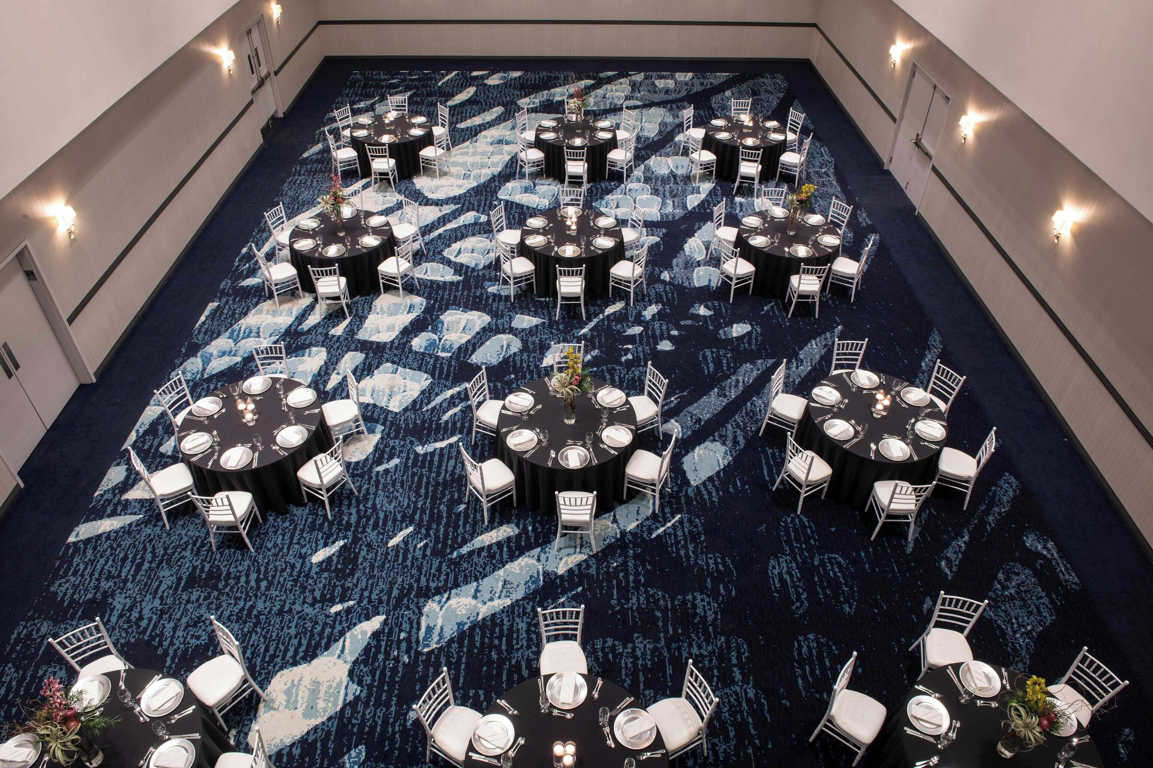 Un amplio salón banqueteado con alfombra, con varias mesas redondas y sillas blancas, dispuestas en un patrón simétrico.