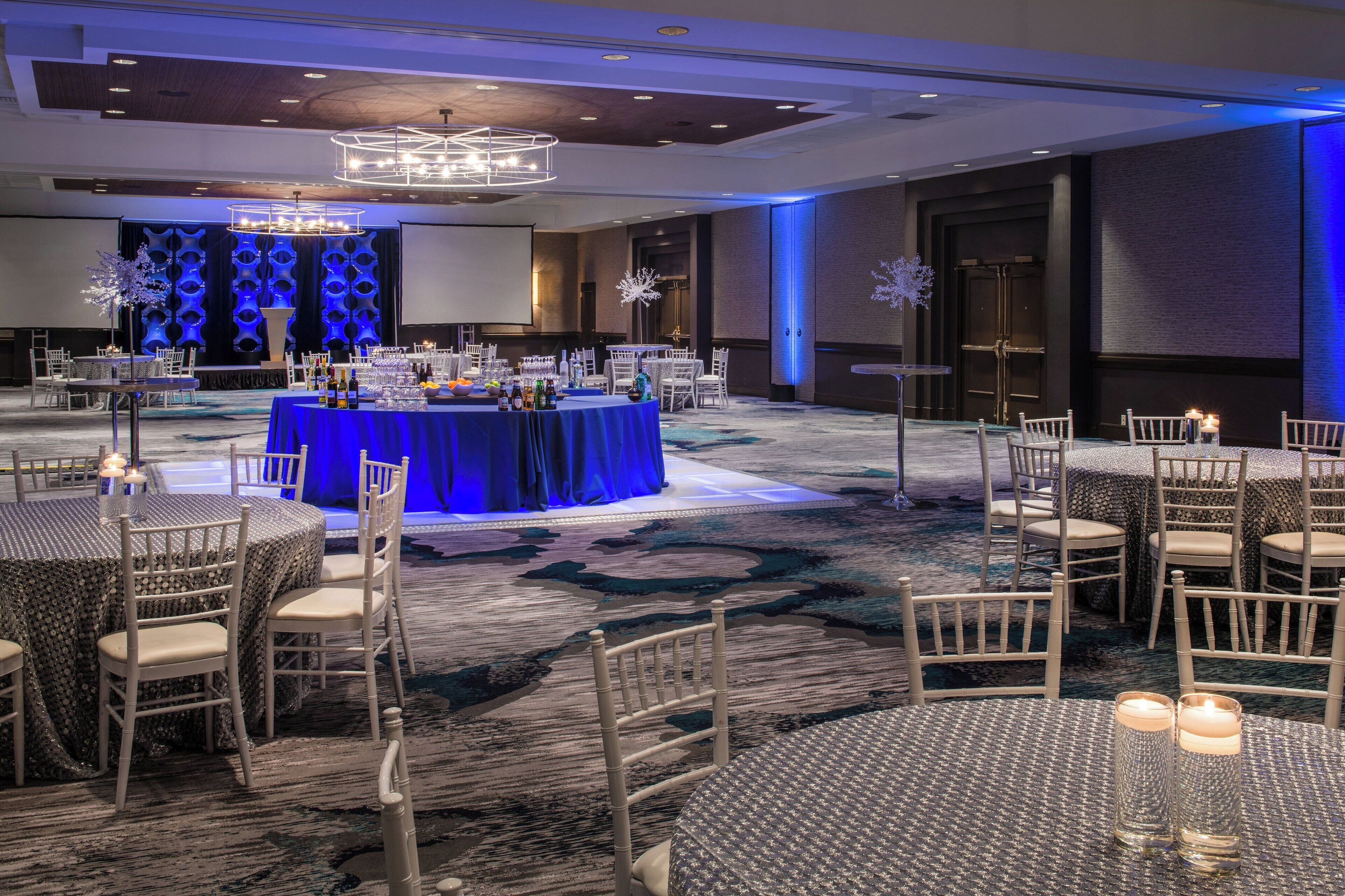 A banquet hall with round tables, chairs, and a stage with a blue backdrop.