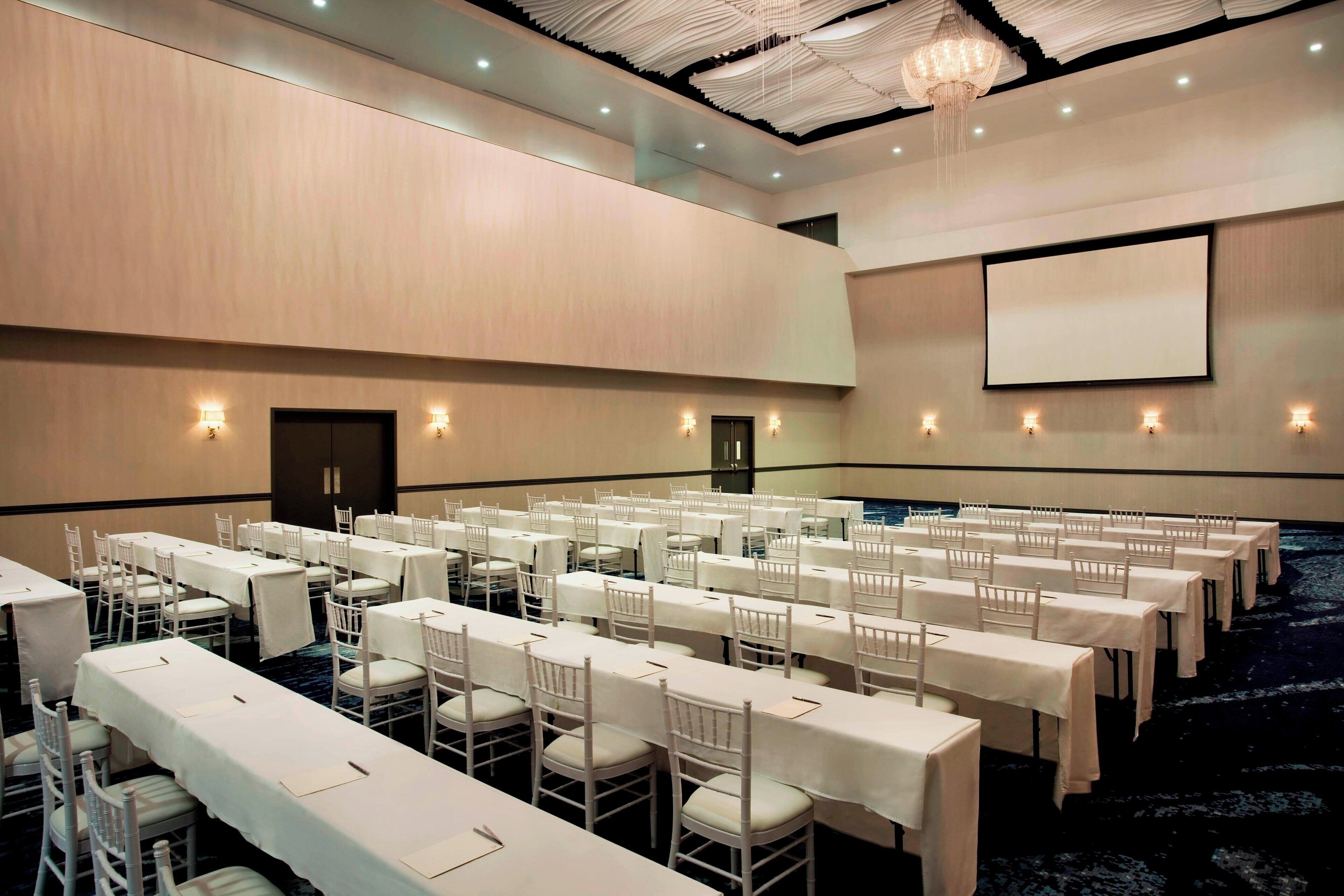 A conference room with multiple tables and chairs, a projector screen, and a chandelier.
