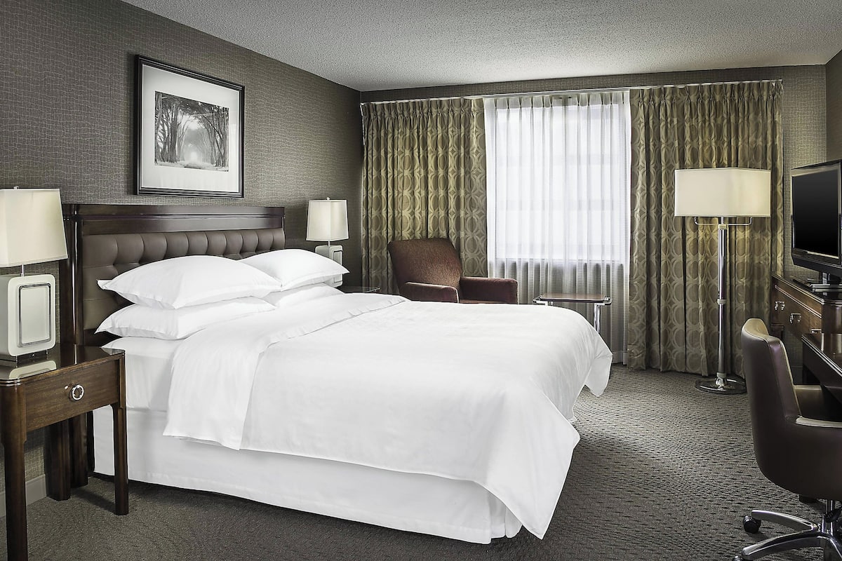 Une chambre d’hôtel avec un grand lit, deux tables de chevet avec des lampes, une chaise, un bureau avec une télévision et une fenêtre avec des rideaux.